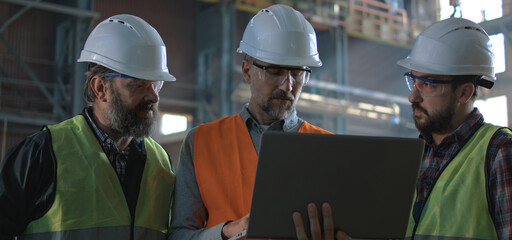 Three professional heavy industry technicians in safety uniform and helmets talk about production. Mature inspector uses laptop standing with colleagues in manufacturing plant or factory. Slow motion.