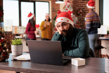 Wall Mural - Exhausted startup company man employee working overtime for project deadline in office at christmas eve. Tired manager in santa hat having burnout in workplace during winter holiday