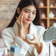 © fitpinkcat84 - Facial beauty skin care, cute asian young woman, girl in bathrobe reflection into mirror, hand applying moisturizer lotion on her face, holding jar of skin cream before makeup cosmetic routine at home