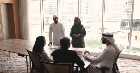 Wall Mural - Two Muslim male and female professionals standing at meeting table, speaking to colleagues, presenting project to European business partners, investors, customers. Diverse team discussing cooperation