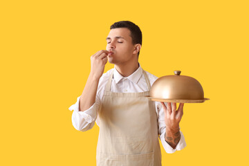 Poster - Handsome happy male chef with tray and cloche showing perfect gesture on yellow background