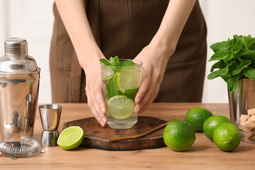 Wall Mural - Young woman with glass of fresh mojito, shaker and measure cup on table in kitchen