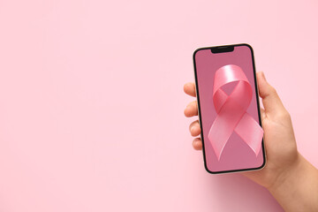 Wall Mural - Female hand with modern mobile phone and pink ribbon on color background, closeup. Breast cancer awareness concept