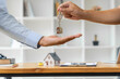 © kudosstudio - Close up hand of a male realtor holding house key chain about to drop on another man's hand, with a small house model on a table