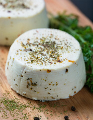 Creamy block of homemade goat cheese with herbs and cracked black pepper on wooden board.