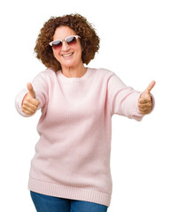 Poster - Beautiful middle ager senior woman wearing pink sweater and sunglasses over isolated background approving doing positive gesture with hand, thumbs up smiling and happy for success