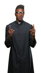 Wall Mural - Young african american priest man over isolated background amazed and surprised looking up and pointing with fingers and raised arms.