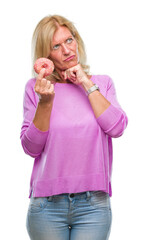 Poster - Middle age blonde woman eating pink donut over isolated background serious face thinking about question, very confused idea