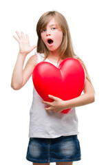 Sticker - Young blonde toddler holding a red heart very happy and excited, winner expression celebrating victory screaming with big smile and raised hands