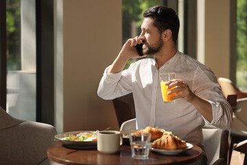 Canvas Print - Happy man having tasty breakfast while talking on smartphone in cafe, space for text