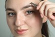 © New Africa - Young woman plucking eyebrow with tweezers on light background, closeup
