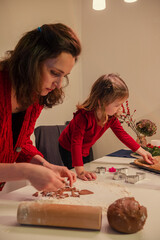 A young mother and her beautiful daughter are making Christmas cookies. The mother and daughter, wearing red sweaters, are making New Year's style cookies, cutting out different shapes with molds