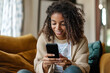 © PrettyStock - Smiling young woman with curly hair using a smartphone on a cozy couch in a relaxed home setting.