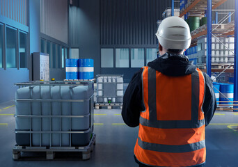 Man is standing in warehouse. Storehouse employee. Warehouse manager has his back to camera storage hangar with fuel in barrels. Chemical products warehouse. Storekeeper in orange vest