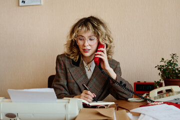 Poster - Portrait of woman speaking on phone while writing down notes at her desk in an office setting with paperwork and supplies scattered around her workspace