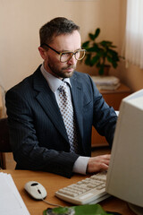 Leinwandbilder - Businessman working on desktop computer in office, wearing suit and glasses while focusing on task, with plant in background adding natural decor
