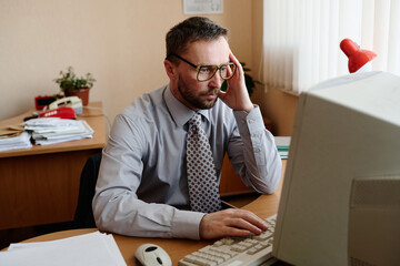 Leinwandbilder - Portrait of a Caucasian man sitting at desk, using a computer in an office environment, wearing glasses and holding head thoughtfully