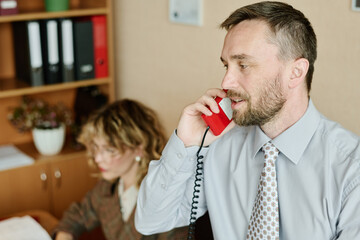 Leinwandbilder - Man in formal attire discussing projects over red wired landline phone while a woman in background focuses on her computer work on desk behind him