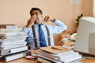 Leinwandbilder - Businessman wearing striped shirt and suspenders yawning and stretching at desk with stacks of paperwork and office equipment around him in corporate environment