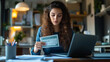 © Maksym - A woman staring blankly at a bill in her hand, her desk cluttered with financial documents and a laptop showing a low bank balance