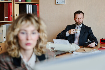 Poster - Man holding coffee cup while sitting at desk, with office supplies and documents scattered around Woman blurred in foreground, creating depth in corporate environment