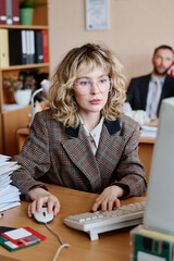 Poster - Portrait of businesswoman with blonde hair looking at the camera while working at her desk with computer, surrounded by office supplies and a colleague in background