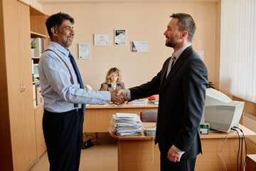 Aufkleber - Two businessmen shaking hands in modern office setting with woman working in background, with documents and computer equipment present on desks creating professional environment