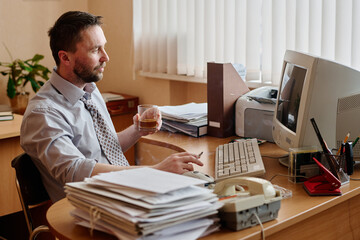 Poster - Caucasian man sitting at wooden desk with glass of beverage in hand, surrounded by office supplies and technology in bright office setting