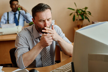 Poster - Man sitting at desk in office, managing stress by smoking while colleague works in background with plants around, maintaining a corporate environment