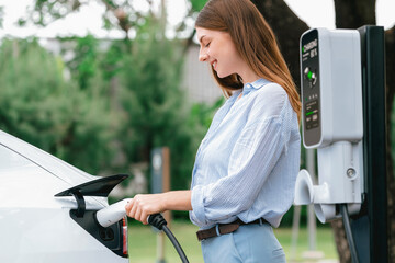 Wall Mural - Young woman recharge EV electric vehicle's battery from EV charging station in outdoor green city park scenic. Eco friendly urban transport and commute with eco friendly EV car travel. Exalt