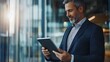 © Esmeralda - A middle-aged Caucasian man with a beard, dressed in a suit, smiles while using a tablet in a modern office with large windows and natural light.