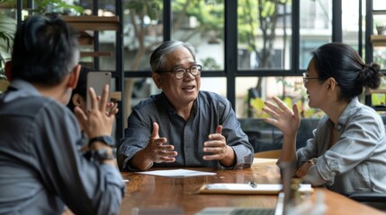 Wall Mural - A group of three colleagues engaged in an animated discussion around a wooden table with large windows and greenery in the background.