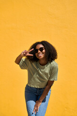 Wall Mural - Happy joyful young African American woman wearing sunglasses having fun looking at camera standing at yellow wall background. Funky gen z Black girl celebrating sunny summer party. Vertical photo.