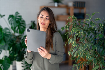 A confident Caucasian female expertly navigates a digital tablet wearing business casual attire, surrounded by indoor foliage in a contemporary office.