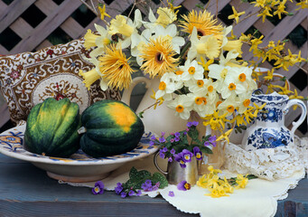 Spring flowers still life of daffodils, narcissi, and gerbera, in vases and jugs on a wooden table with a wooden lattice fence in the background, with two pumpkins in a bowl outdoors