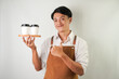 © Rizky - Excited young asian man wearing rolled-up white sleeve shirt and brown apron is holding and serving coffee drinks to customer with smiling face expression. Isolated over white background.