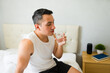 © AntonioDiaz - Young man is sitting on his bed in his bedroom, drinking a glass of water after waking up in the morning