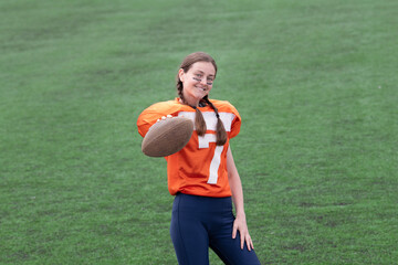 A smiling girl or young woman in a sports uniform is holding a ball in front of her, looking at you, and inviting you to play American football. In the background, there is green grass on the football