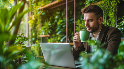 A businessman takes a sip of coffee while focused on his laptop in a green office environment