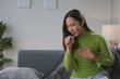 © amnaj - Young asian woman is sitting on a sofa at home, holding her chest and coughing