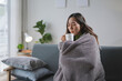 © amnaj - Young woman sitting on a sofa covered with a blanket and holding a mug, warming herself up in the morning