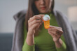 © amnaj - Young woman sitting on a couch at home feeling ill and opening a bottle of pills