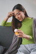 © amnaj - Young woman is sitting on her couch covered with a blanket, holding a pill bottle and feeling sick