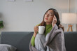 © amnaj - Young asian woman having a headache and holding a mug while sitting on a sofa at home, covered with a blanket