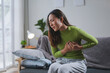 © amnaj - Young woman is suffering from chest pain while sitting on her living room sofa