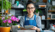 © missisya - A smiling woman in an apron stands in a colorful plant shop, showcasing vibrant flowers and greenery with a tablet in her hands.