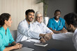 © fizkes - Young Indian male doctor and colleague shaking hands, making gesture of agreement, cooperation, or conclusion of successful discussion, sit at table in modern conference room. Partnership, teamwork
