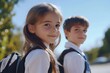 © kinara art design - A young school girl in a white shirt and a black backpack with her long hair down, smiles while standing outdoors with a boy in the background wearing a white shirt and a black backpack.