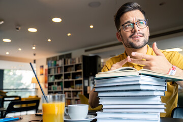 Wall Mural - A young man leans on a stack of books, displaying contemplation, with coffee and juice nearby, in a well-lit library setting.