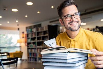 Wall Mural - A cheerful caucasian male student relaxes in a library, with coffee beside him as he leans on a tower of books, ready to study.